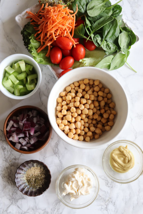 Chickpeas in a white bowl next to carrots, tomatoes, celery, red onions, mustard, mayonnaise, and spices by Naturally Tessa