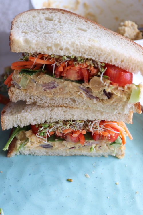 Chickpea salad sandwich on top of a blue plate with tomatoes, carrots, onions, celery and sprouts by Naturally Tessa.